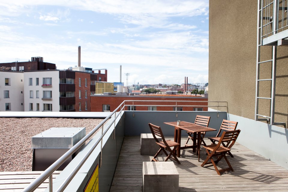 Wooden dining table and four chairs in a wide roof terrace. View is such that you can see far from here. Closer to the terrace, the neighbouring buildings' roofs are seen.